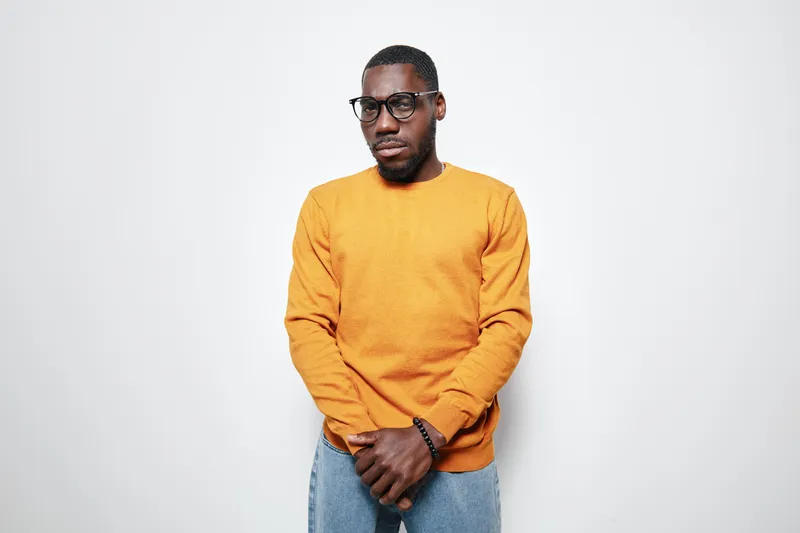 African American man with dark skin wearing glasses and orange sweater standing against white background looking thoughtful and serious in casual style portrait.