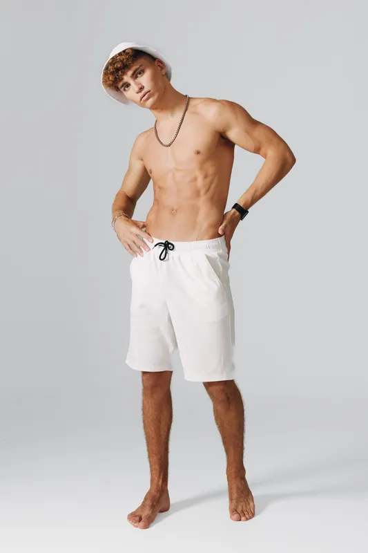 Young man posing shirtless with white shorts and chain necklace, wearing hat and smart bracelet on clean background, barefoot and casual standing for fitness and style concepts.