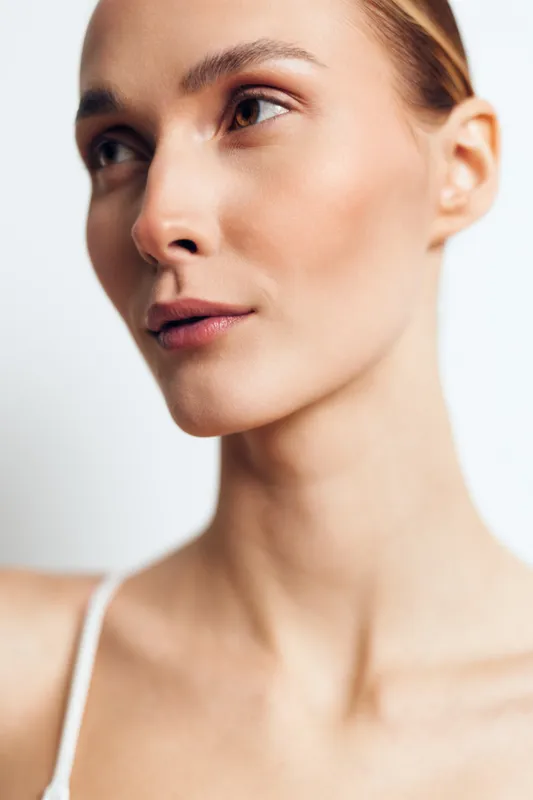 Close-up portrait of a young woman with clear skin looking away. Soft natural lighting highlights her smooth complexion and delicate facial features on a plain background.