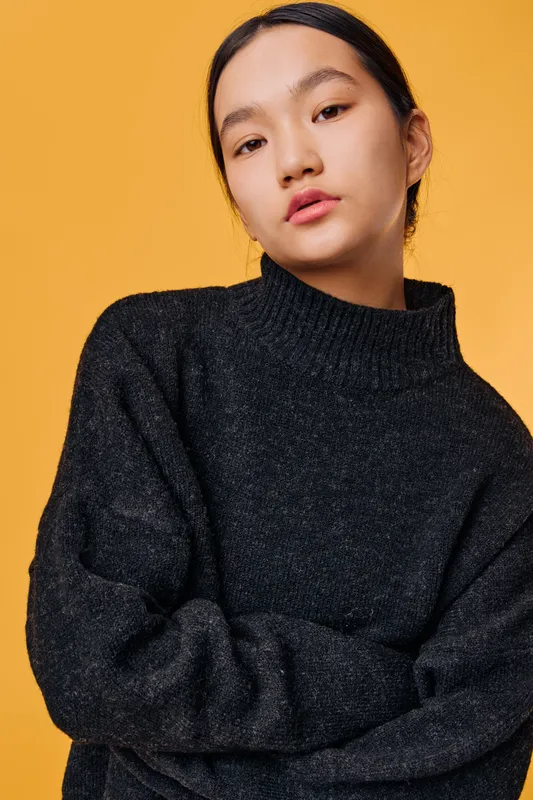 Young woman with dark hair wearing a black sweater posing with folded arms against a yellow background in a studio. Portrait of confident female model with natural makeup and casual outfit.