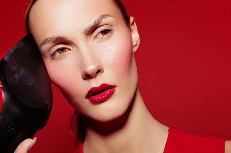 Close-up of a woman with red lipstick holding a black high heel shoe near her face on a red background. Concept of fashion, beauty, and style with bold colors.