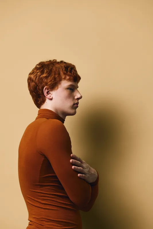 Young man with red hair and pale skin wearing brown turtleneck sweater standing sideways with hands crossed on light beige background studio portrait.