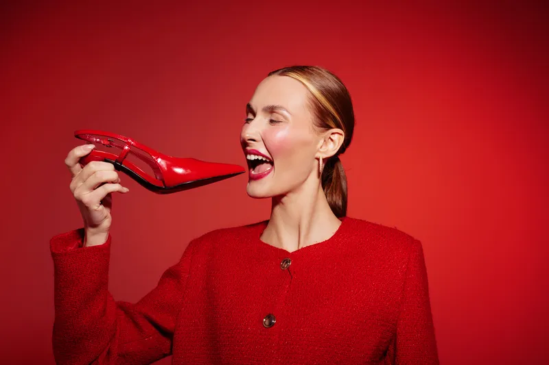 Woman in red jacket holds red high heel shoe near face with eyes closed and smiling. Studio shot with red background. Concept of fashion, style, and joyful expression.