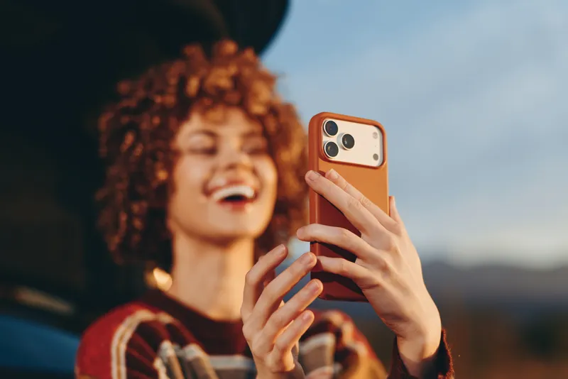 Woman selfie smartphone lifestyle outdoors portrait sunset closeup, young adult smiling, curly hair and freckles, holding phone for camera, warm golden hour light, blurred mountains background