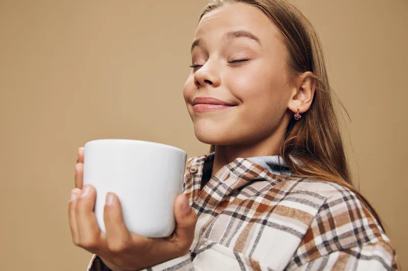 Girl holding a large white cup close to her face with closed eyes, smiling with satisfaction and wearing a plaid shirt against a plain beige background in studio.
