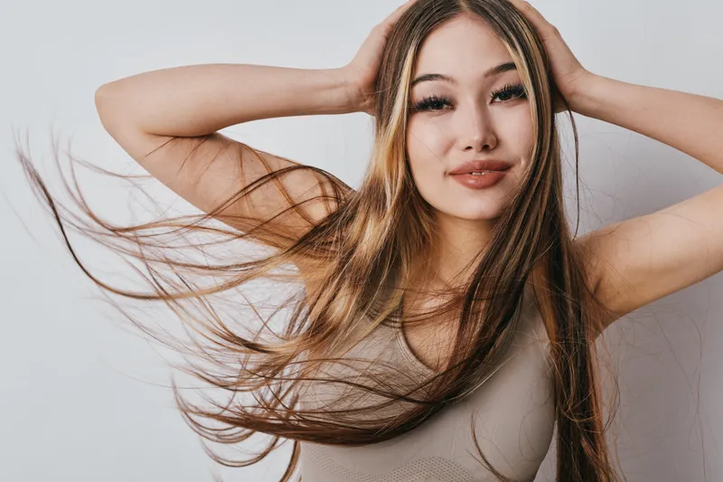 Young woman with long flowing hair poses with hands behind head against white background, smiling gently in casual tank top fashion portrait.