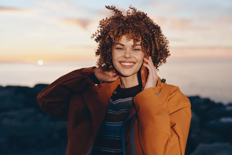 Woman lifestyle outdoor sunset portrait, smiling with curly hair, wearing a brown jacket, relaxed pose, warm evening light, coastal landscape behind, candid travel mood at dusk.