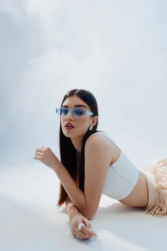 Young woman in white top wearing blue sunglasses lies on white surface. Her long dark hair is styled straight. Concept of beauty, fashion, and modern style.