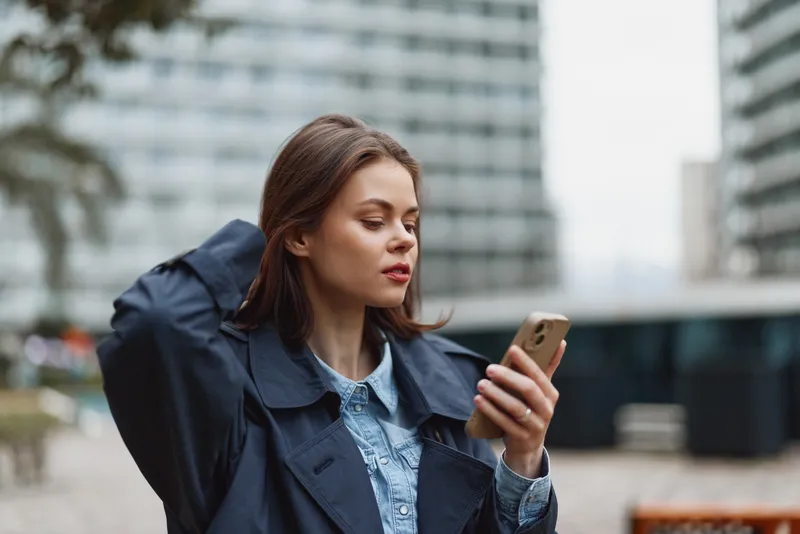 Woman outdoors wearing denim jacket, focused on phone, urban city backdrop, casual moment, thoughtful expression, fashion forward vibe, gentle breeze, natural colors, soft light, neutral tones