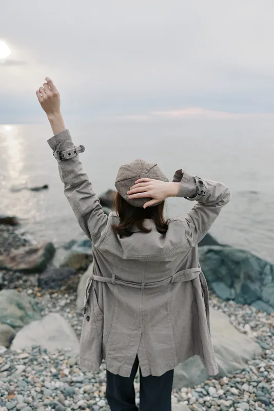 Woman in checkered coat and hat stands on rocky beach with arms raised, looking at calm sea under cloudy sky. Concept of freedom, relaxation, and nature connection.