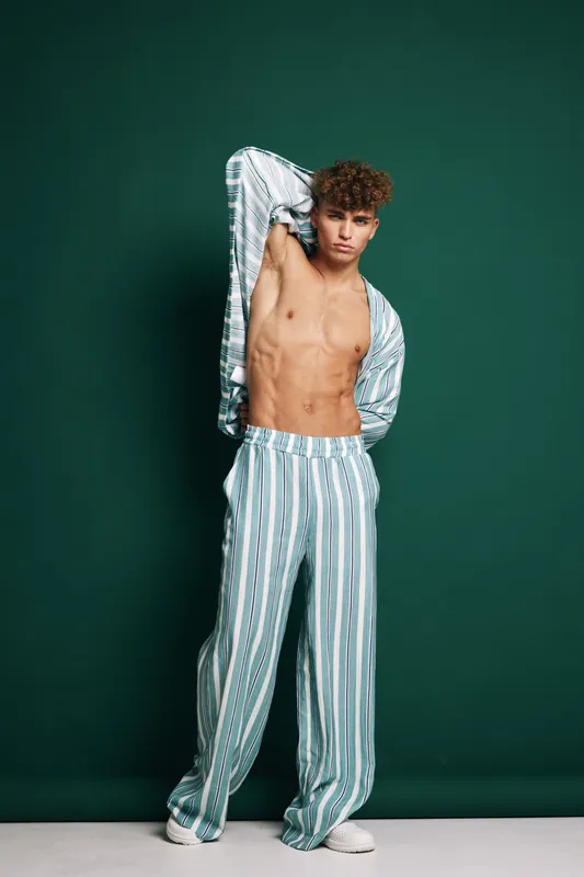 Young man with curly hair wearing striped pajamas posing against dark green background, showing toned body and relaxed confident expression in studio setting.