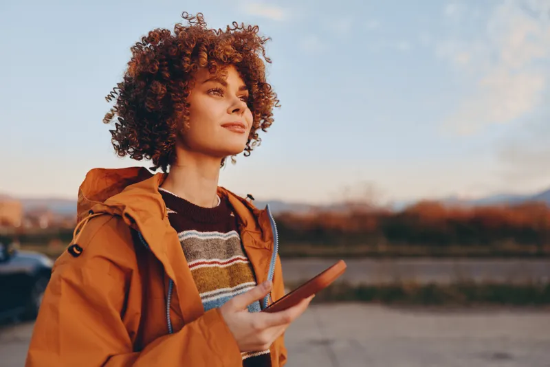 Woman lifestyle outdoors with curly hair and orange jacket, holding smartphone by a river at sunset, relaxed portrait with natural light, casual travel mood, evening breeze
