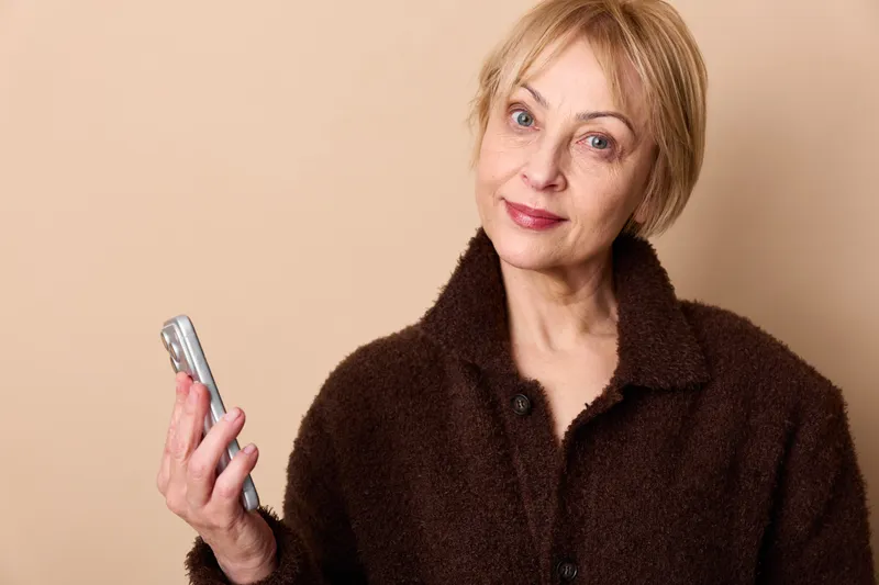 Mature woman with smartphone in hand, indoor portrait, gentle smile, holding phone close to face, calm communication with technology device, soft beige background