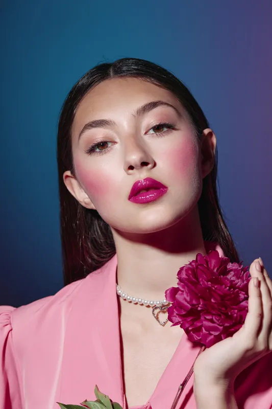 Fashion model with glossy pink lips and vibrant blush poses holding deep magenta flower. Creative beauty portrait with pearl necklace on solid blue background.
