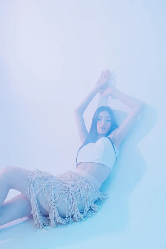 Young woman beauty fashion model poses with arms raised wearing a white top and fringed skirt. Soft pastel lighting creates a dreamy studio atmosphere for style concept.