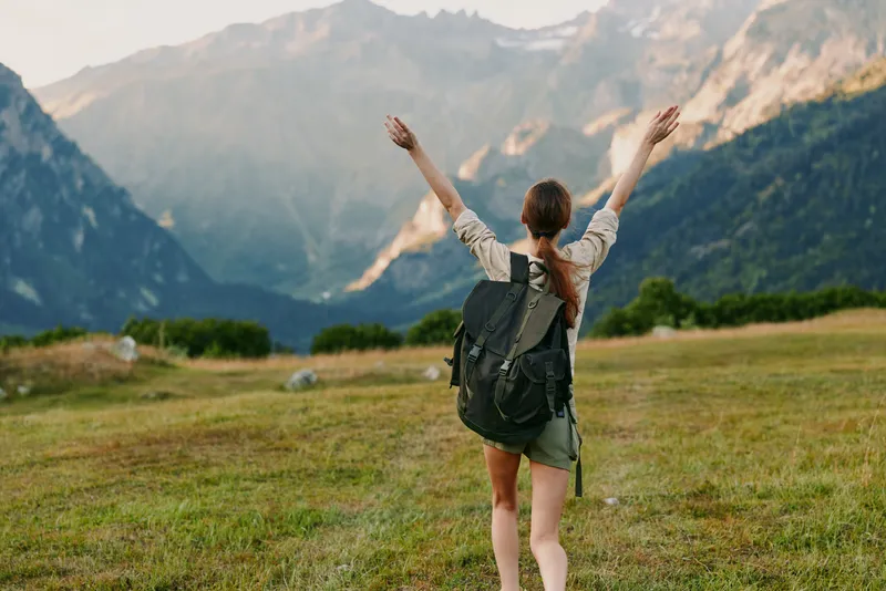 Young woman traveler with backpack raises hands enjoying mountain landscape on sunny day, standing on green grass field with clear sky and distant peaks.