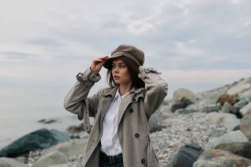 Young woman in a gray coat and hat stands on a rocky beach looking into the distance under cloudy sky. Concept of autumn fashion, outdoor lifestyle, and thoughtful mood.
