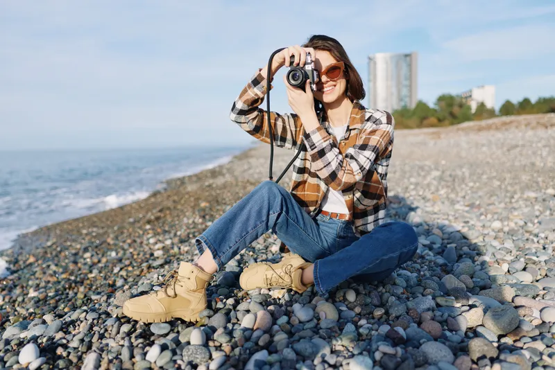 Photographer on a pebble beach captures with a camera, enjoying travel and outdoor lifestyle, a casual seaside portrait under daylight with sunglasses and boots, expressing relaxation and exploration