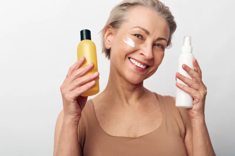 Smiling mature woman holding two skincare bottles with cream on her cheek, posing on white background. Concept of beauty routine, facial care, and natural skincare products.