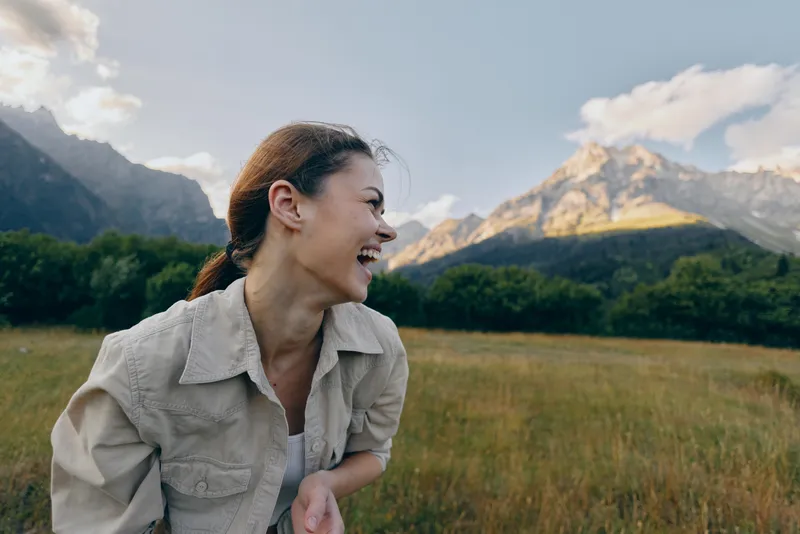 Young woman laughs in an open meadow with mountains behind, casual jacket, sunlight and breeze creating a relaxed lifestyle moment