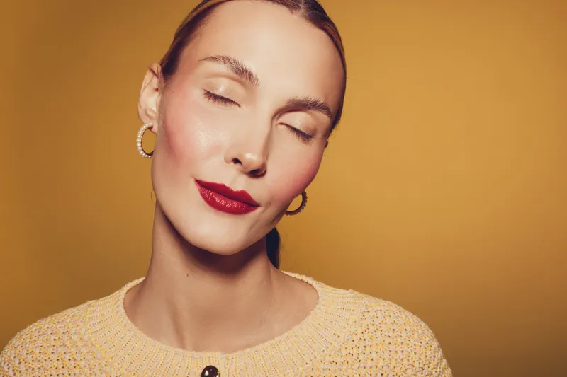 Close-up portrait of a woman with closed eyes wearing red lipstick and gold hoop earrings. Soft lighting highlights her smooth skin and calm expression on a warm beige background.