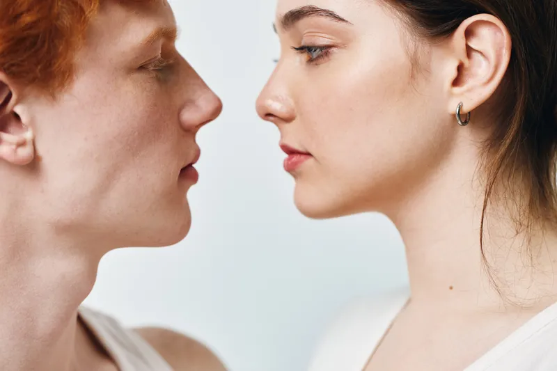 Young man and woman facing each other in closeup portrait with neutral expression on white background. Skin details and side profiles in intimate minimal composition for relationships.