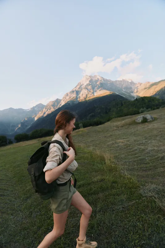 Young woman with a backpack hikes through a grassy mountain meadow during golden hour, enjoying the peaceful natural landscape and fresh air with a relaxed expression.