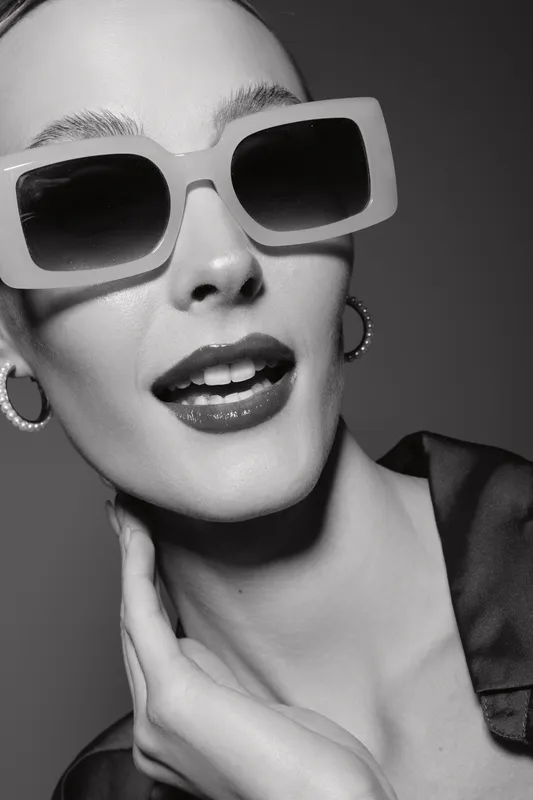 Close-up portrait of a young white woman wearing large sunglasses and hoop earrings, posing confidently with hand near neck in black and white.