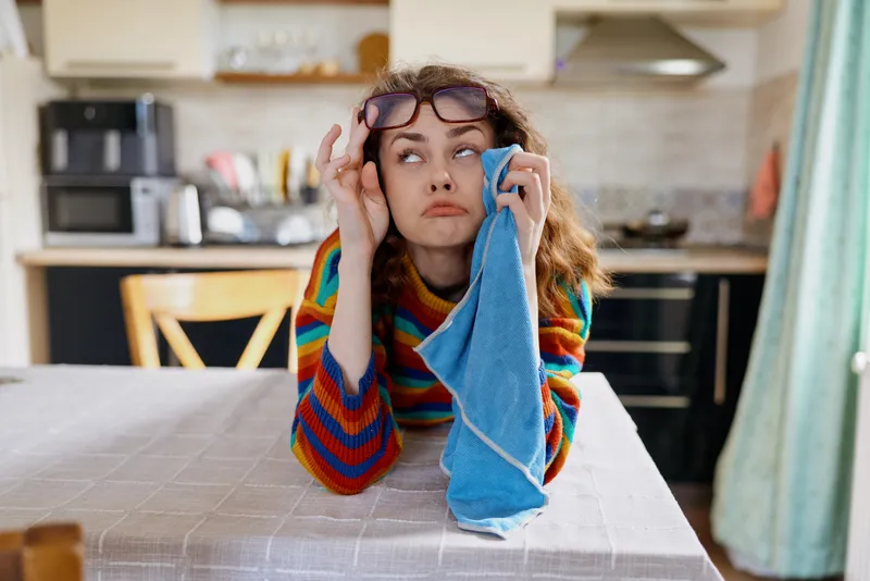 Woman in a colorful striped sweater sits at a home kitchen table, holding a blue cloth to her face with glasses, showing tired mood during a domestic cleaning session, reflecting everyday home life