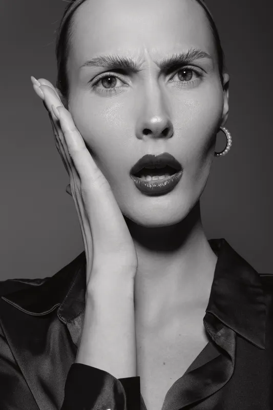 Close-up of a surprised Caucasian woman with fair skin and pulled-back hair, wearing hoop earrings and a black shirt, posing against a grey background.