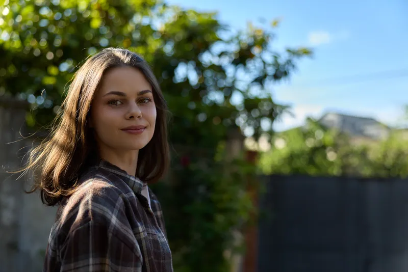 Smiling woman in a plaid shirt outdoors in a sunny garden. A lifestyle portrait focused on casual style, nature, well being, and confident mood under warm daylight