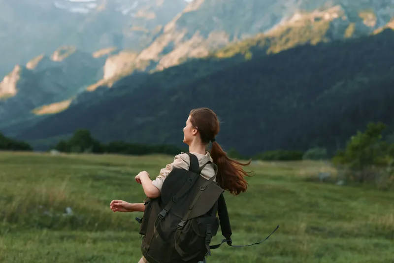 Woman hiker with backpack crossing a grassy meadow beneath distant mountains, capturing an outdoor exploration moment with freedom and a bright, natural light mood.