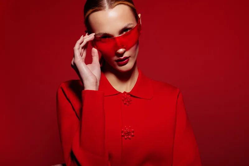Stylish woman in red sweater and heart-shaped sunglasses poses against red background. Fashion portrait with bold color contrast and modern accessories concept.