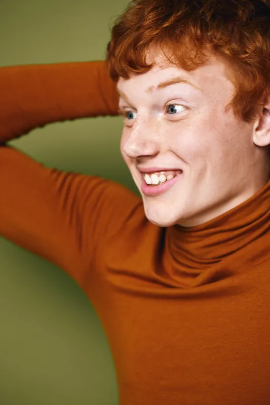 Young man with red hair wearing brown turtleneck smiling looking sideways with hand behind head against green background. Emotion, casual, portrait, happiness, cheerful young adult.