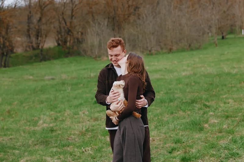 A smiling couple in casual autumn clothing embrace in a green park, with a small dog resting in their arms. A candid lifestyle moment capturing warmth, connection, and outdoor happiness.