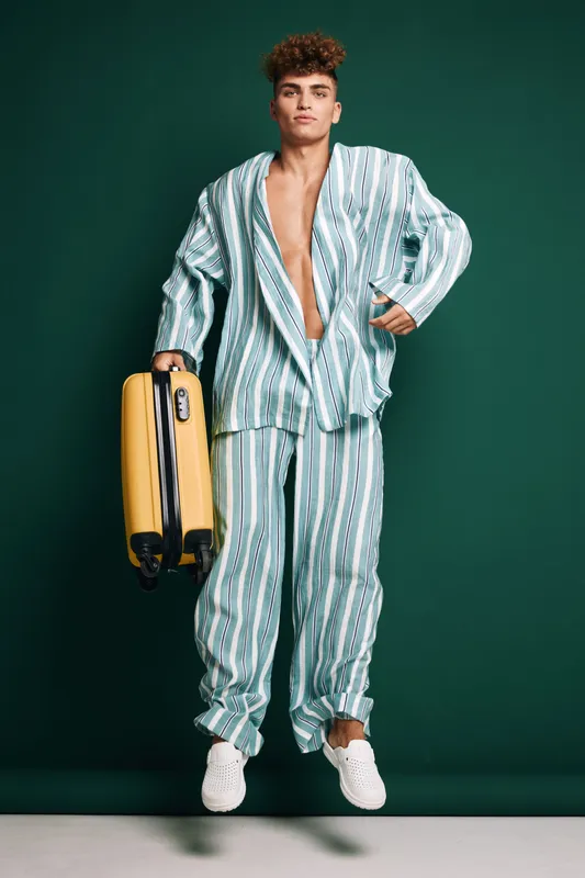 Young man in striped pajamas holding yellow suitcase standing against green background with casual white shoes and confident expression in studio.