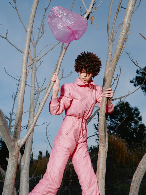 Person in pink jumpsuit holding a pink balloon among bare trees, outdoor portrait with whimsical fashion and playful mood under sunny natural daylight.