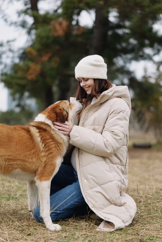 Dog, woman, coat, winter, outdoors, interaction, affection, pet, friend, park, a woman kneeling in a warm coat and beanie pets a friendly dog in an open grassy park.