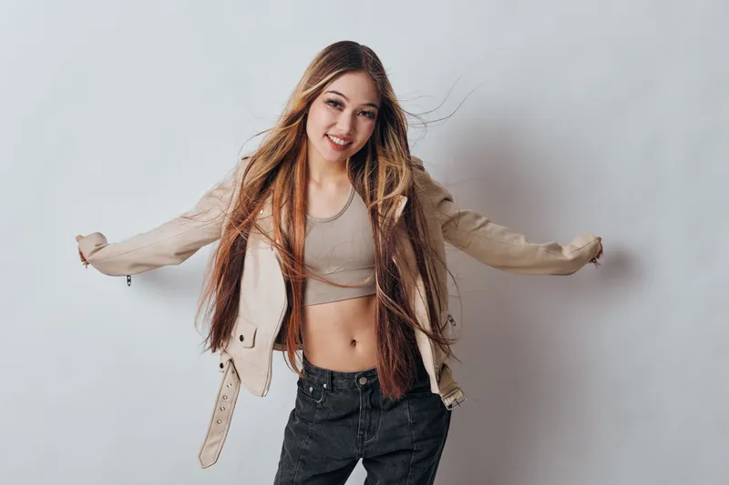 Young woman with long hair smiling and posing in casual outfit with crop top and jacket against gray background, expressing joy and confidence with open arms.