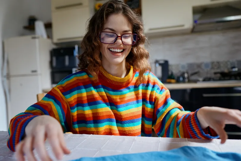 Smiling woman with curly hair wearing a colorful striped sweater sits at a kitchen table, engaging in a creative home project, bright natural light, cozy domestic mood, candid
