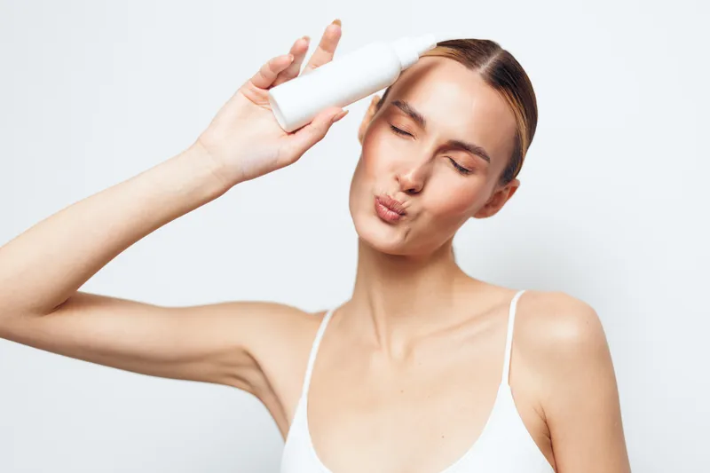 Young woman with slicked back hair holds a white cosmetic bottle near her face while making a playful kiss expression against a plain white background.