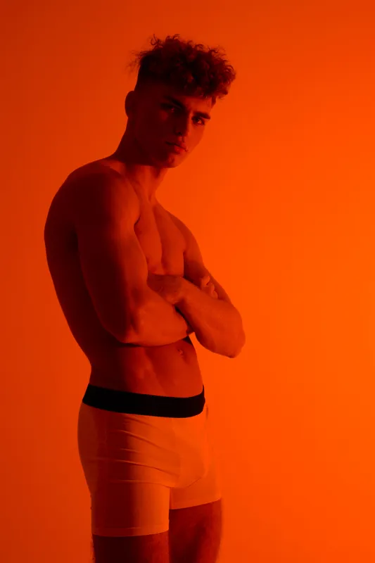 Man with muscular body and curly hair posed with arms crossed wearing white underwear with black stripe. Orange ambient lighting and fitness male model in studio portrait.