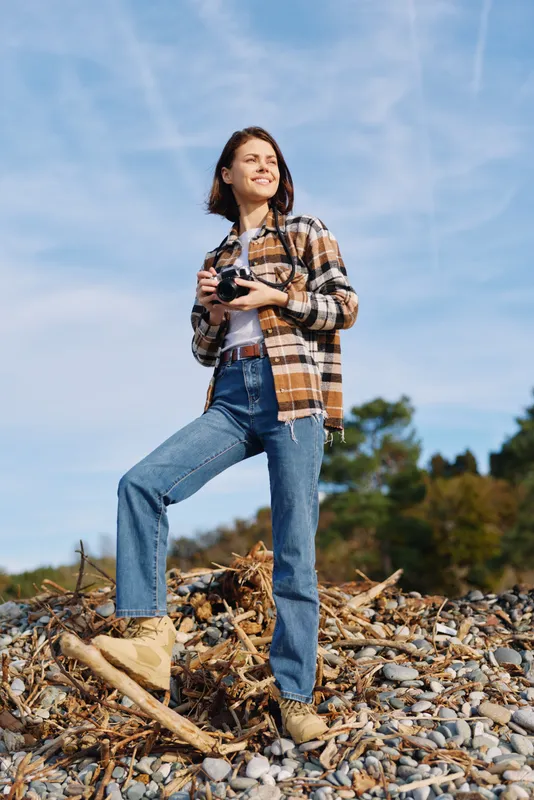 Woman photographer standing on a rocky shore with camera, wearing a plaid shirt and jeans, gazing up at a bright blue sky, wind in her hair, capturing outdoor landscape moments.