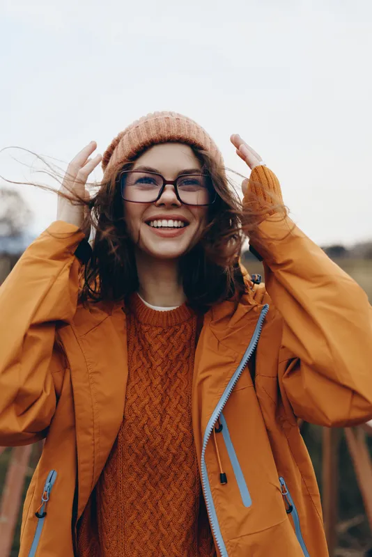 Woman lifestyle outdoors autumn smiling glasses hat coat portrait hands pose, woman adjusts knit hat with both hands, orange jacket, blurred background, warm daylight, casual street