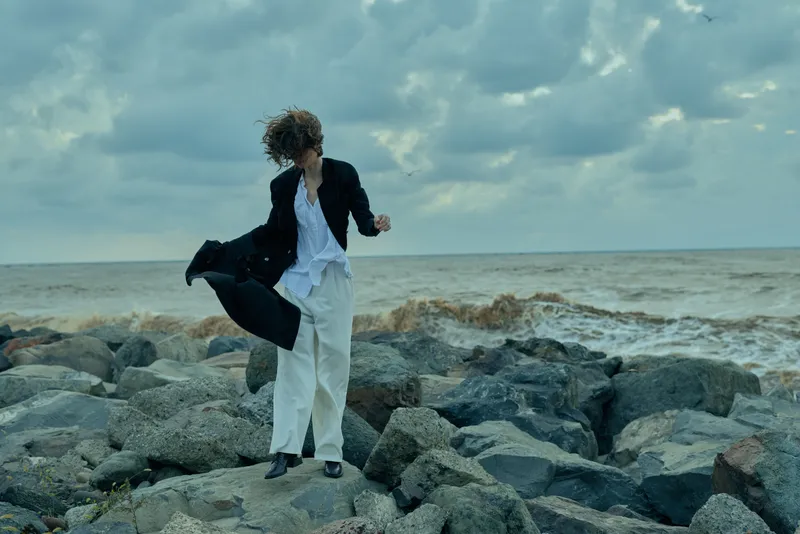 Coastal model balancing on rugged rocks by the sea, windblown coat and white outfit, moody sky and dynamic pose capture fashion against nature's edge, emphasizing movement and style in a dramatic