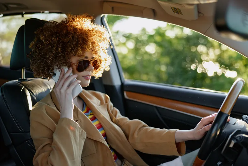 Cheerful woman driving a car with retro film color, sunlit interior, curly hair, sunglasses, blazer, relaxed smile, casual vibe, outdoorsy moment, warm tones and motion
