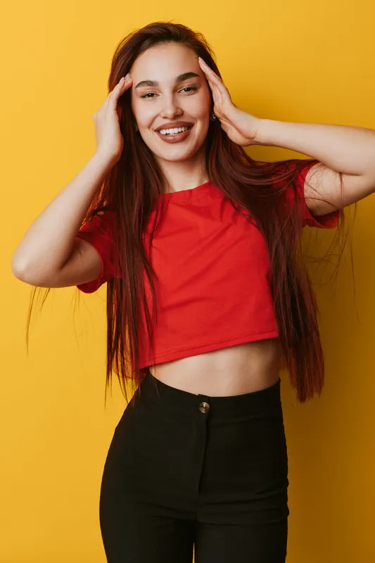 Young woman with long hair wearing red crop top and black pants posing with hands near head smiling on yellow background. Casual fashion, beauty and happiness concept.