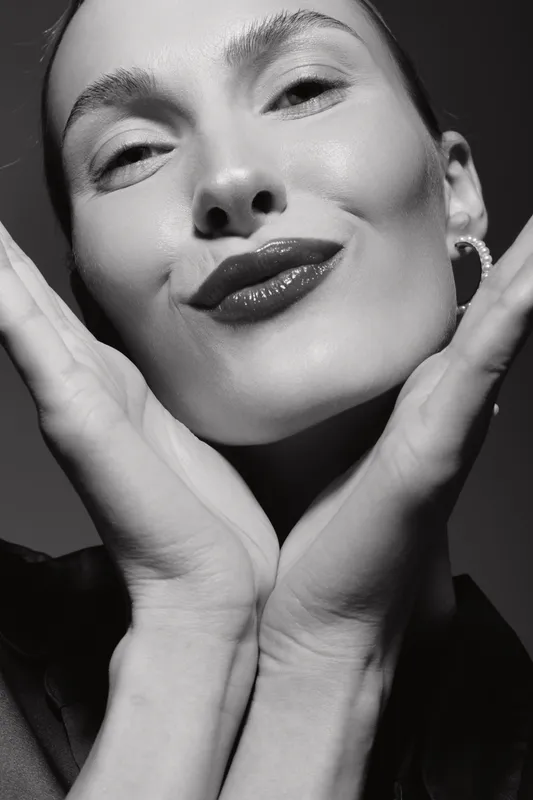Close-up portrait of a fair skinned white woman with glossy lips and earrings making a playful face with hands near her cheeks.