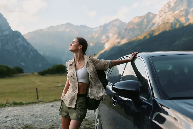 Young woman leans against a car in a rugged mountain landscape, enjoying fresh air under clear skies with a relaxed smile and casual outfit