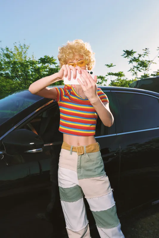 Woman lifestyle retro fashion outdoors beside a car wearing striped patchwork pants, she covers her face with pink fabric, cheerful playful mood, bright daylight, vintage film color